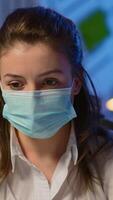 Vertical . Close up of woman with face mask reading emails late at night to respect deadline of project working in new normal business office, taking notes, analysing documents overtime during global pandemic video