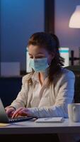 Vertical . Manager woman with face mask working overtime in new normal business office checking team project, taking notes, analysing financial documents sitting at desk late at night during global pandemic video