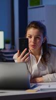 Vertical . Business woman discussing during conference with partners using smartphone and headphone late at night working in start-up office. Employee with modern technology network wireless doing overtime video
