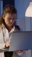Vertical . Focused businesswoman checking business reports standing in start up office late at night typing on laptop. Busy employee using technology network wireless doing overtime reading writing, searching video