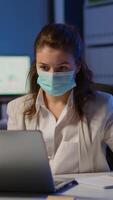 Vertical . Employee with protection face mask working late at night in new normal business office to respect deadline of project, taking notes, analysing documents sitting at desk overtime during global pandemic video