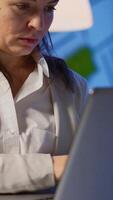 Vertical . Close up of concentrated entrepreneur analysing financial reports standing in start up office late night holding laptop. Focused employee using technology network wireless overtime reading, searching video