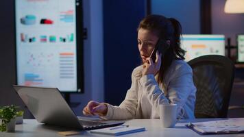 Woman employee speaking at phone while working at laptop late at night. Busy focused freelancer using modern technology network wireless doing overtime for job reading writing, searching taking break video
