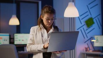 Focused businesswoman checking business reports standing in start up office late at night typing on laptop. Busy employee using technology network wireless doing overtime reading writing, searching video