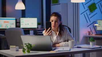 Business woman discussing during conference with partners using smartphone and headphone late at night working in start-up office. Employee with modern technology network wireless doing overtime video