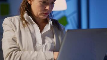Close up of concentrated entrepreneur analysing financial reports standing in start up office late night holding laptop. Focused employee using technology network wireless overtime reading, searching video