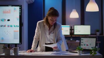 Stressed manager woman working with financial documents checking graphs, holding papers, reading raports, standing at desk late at night in start-up office doing overtime to respect deadline. video