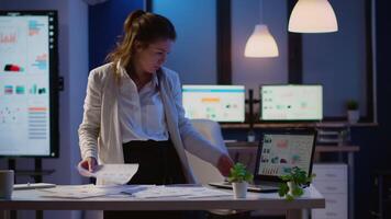 Focused manager woman checking business reports standing in start up office late at night holding laptop. Busy employee using technology network wireless doing overtime reading writing, searching video