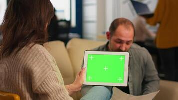 Over the sholder of manager woman sitting on couch talking with coworker holding tablet with green screen while diverse team working. Multiethnic people planning project on chroma key display video