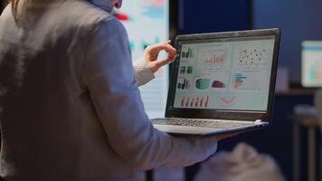 Back view of business woman analysing financial statistics holding laptop standing in start-up office late at night overworking. Busy focused employee using modern technology network wireless overtime video