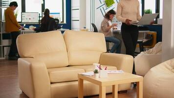 Focused black manager reading tasks on laptop and typing sitting on couch in busy start up office while diverse team analyzes statistics data in background. Multiethnic team talking about project video