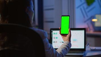 mulher empregado olhando às Smartphone com croma chave sentado às escrivaninha dentro começar acima o negócio escritório atrasado às noite. o negócio mulher assistindo Área de Trabalho exibição com verde brincar, tela, trabalhando ao longo do tempo. video
