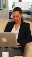 Hispanic business woman smiling at camera sitting on couch typing on computer while diverse colleagues working in background. Multiethnic coworkers analysing startup financial reports in modern office video