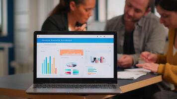 Laptop screen ready for financial project presentation placed on desk while business people work in background. Diverse employees gathered in co-working, working process in busy company, teamwork help video