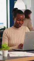 Happy black manager reading tasks on laptop and typing sitting at desk in busy start up office while diverse team analyzes statistics data in background. Multiethnic group talking about project video