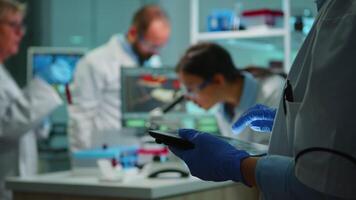 Close up of chemist nurse using tablet while team of scientists doing biological researches under microscope in backgorund late at night. Writing data into notepad in modern equipped laboratory. video