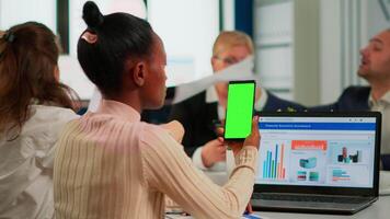 Back view of black businesswoman sitting at conference desk holding smartphone with green screen while colleagues working on background. Multiethnic coworkers planning project on chroma key display video