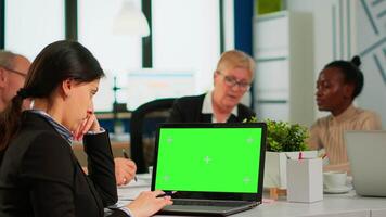 Over the sholder of manager sitting at meeting desk looking at laptop with green screen mockup while diverse team working on background. Multiethnic people planning project on chroma key display video