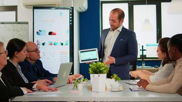 Focused executive manager presenting annual financial report holding tablet standing at conference table in front of diverse businesspeople. Multiethnic team working in startup company video