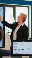 Elderly project manager pointing at desktop presenting statistical data, briefing diverse group of employees. Multiethnical team working in professional startup financial company during conference video