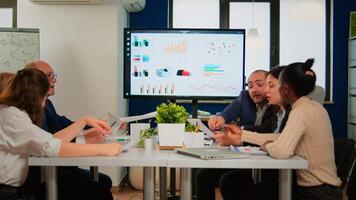 Man leader pointing at digital interactive whiteboard sitting at brainstorming table discussing with coworkers in meeting room. Executive explaining company's vision in broadroom with TV screen video