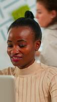 Portrait of delighted african business woman reading good news on laptop sitting at desk in busy start up office while diverse team analyzes statistics data. Multiethnic team working at new project video