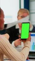Back view of black businesswoman sitting at conference desk holding smartphone with green screen while colleagues working on background. Multiethnic coworkers planning project on chroma key display video