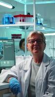 Vertical . Portrait of senior scientist woman looking exhausted at camera sitting in equipped lab late at night. Team examining virus evolution using high tech for scientific research, vaccine development video