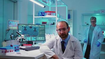 Portrait of experienced scientist man looking tired at camera working overtime in equipped lab. Examining virus evolution using high tech and chemistry tools for vaccine development. video