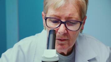 Close up of senior scientist making research using microscope while colleagues working on background in modern equipped laboratory. Stuff analysing vaccine evolution using high tech video