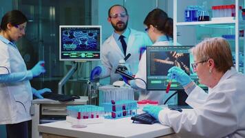 Senior chemist analysing blood sample while coworkers discussing in background about vaccine development working overtime in chemistry equipped laboratory. video