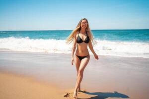 Smiling woman in bikini walking on sandy beach along sea waves photo