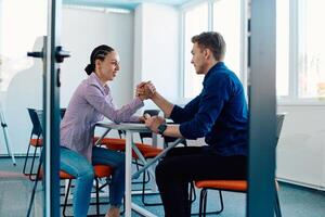 In a modern startup office, a businessman and a businesswoman business colleagues engage in a symbolic arm-wrestling match, reflecting teamwork, competition, and innovation in their dynamic workplace. photo
