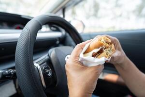 Asian lady holding hamburger and French fries to eat in car, dangerous and risk an accident. photo