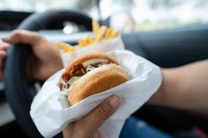 Asian lady holding hamburger to eat in car, dangerous and risk an accident. photo