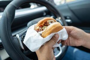 Asian lady holding hamburger to eat in car, dangerous and risk an accident. photo