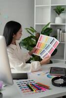 Asian woman freelance graphic designer working with color swatch samples and computer at desk in home office, young lady choosing color gamma for new design project photo