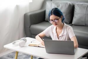 Happy young woman asian using laptop sitting at desk writing notes while watching webinar, studying online, looking at laptop screen learning web classes or having call remote from home photo