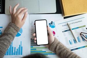 Top view of business woman using smartphone with blank white screen and laptop computer for accounting and financial photo