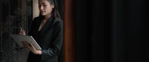 Attractive business asian woman using a digital tablet while standing in front of windows in an office building photo
