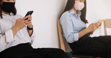 dos asiático joven mujer chateando en teléfono inteligente y vistiendo máscara sentado distancia proteger desde covid-19 virus para social distanciamiento para infección riesgo. foto