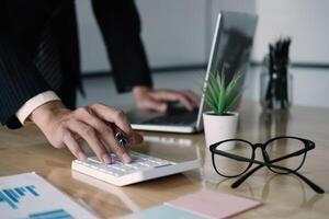 Close up of businessman or accountant hand holding pen working on calculator to calculate business data, accountancy document and laptop computer at office, business concept photo