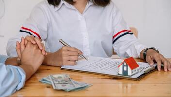 Cropped image of real estate agent assisting client to sign contract paper at desk with house model photo