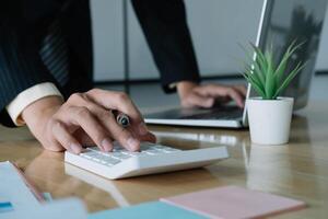 Close up of businessman or accountant hand holding pen working on calculator to calculate business data, accountancy document and laptop computer at office, business concept photo