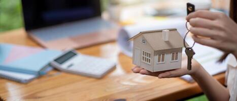 mortgage, real estate and property concept - close up of hands holding house model and home keys with financial working space background. photo