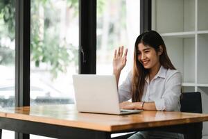 Smiling asian girl talking and waving hand hello during online call via laptop computer, sitting in coffee shop. Cheerful business woman having conference in internet via portable notebook. photo