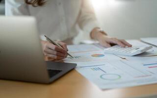 Close up of businesswoman or accountant hand holding pen working on calculator to calculate business data, accountancy document and laptop computer at office, business concept. photo