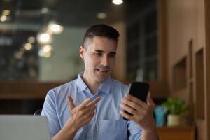 Cheerful happy businessman using smartphone app for virtual communication, holding phone in hand and waving at webcam, holding call sitting at the desk. photo