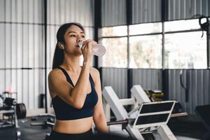asiático joven mujer Bebiendo agua después rutina de ejercicio en gimnasia, saludable y deporte concepto. foto