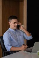 Portrait of young smiling cheerful entrepreneur in modern office making phone call while working with laptop. photo